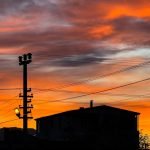 A vibrant sunset over silhouettes of rooftops and power lines, creating a dramatic sky scene.