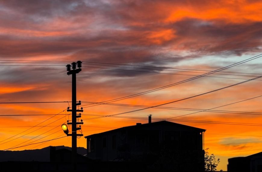 A vibrant sunset over silhouettes of rooftops and power lines, creating a dramatic sky scene.