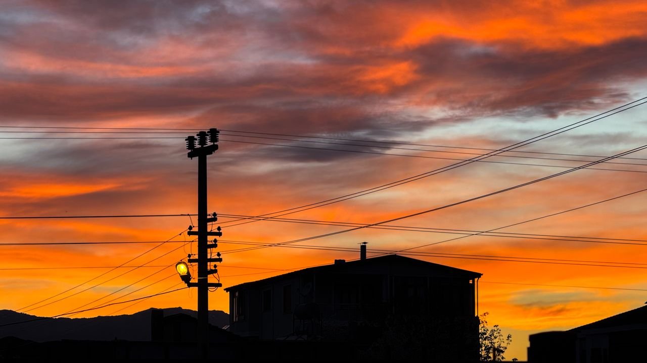 A vibrant sunset over silhouettes of rooftops and power lines, creating a dramatic sky scene.