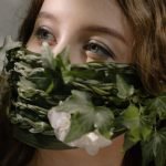 A woman's face adorned with a creative mask made of green leaves and flowers.