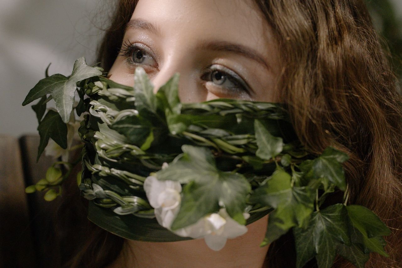A woman's face adorned with a creative mask made of green leaves and flowers.