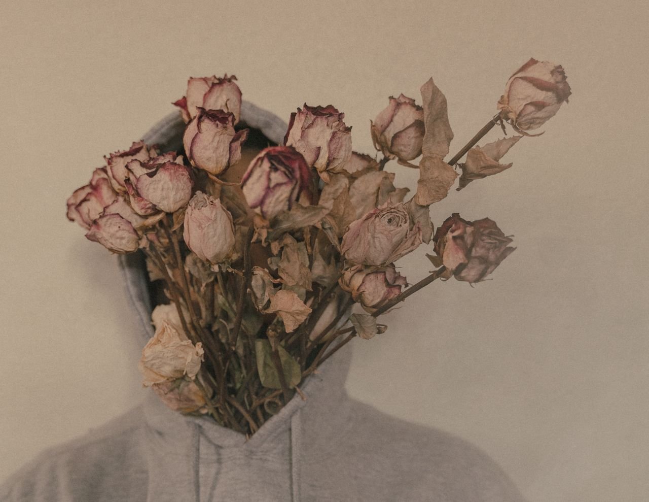 Artistic photo of a person with a bouquet of withered roses covering their face, wearing a hoodie.