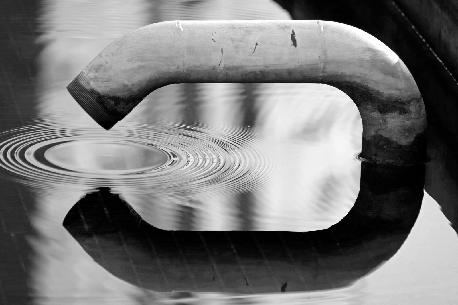 Black and white image of a pipe creating ripples in water. An abstract reflection scene.