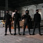 Four masked individuals stand in a dark, empty industrial space, creating an intense atmosphere.