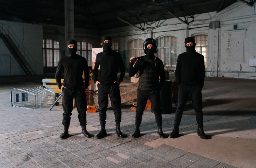 Four masked individuals stand in a dark, empty industrial space, creating an intense atmosphere.