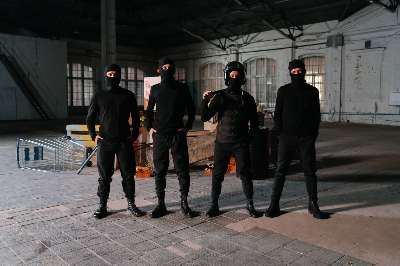 Four masked individuals stand in a dark, empty industrial space, creating an intense atmosphere.