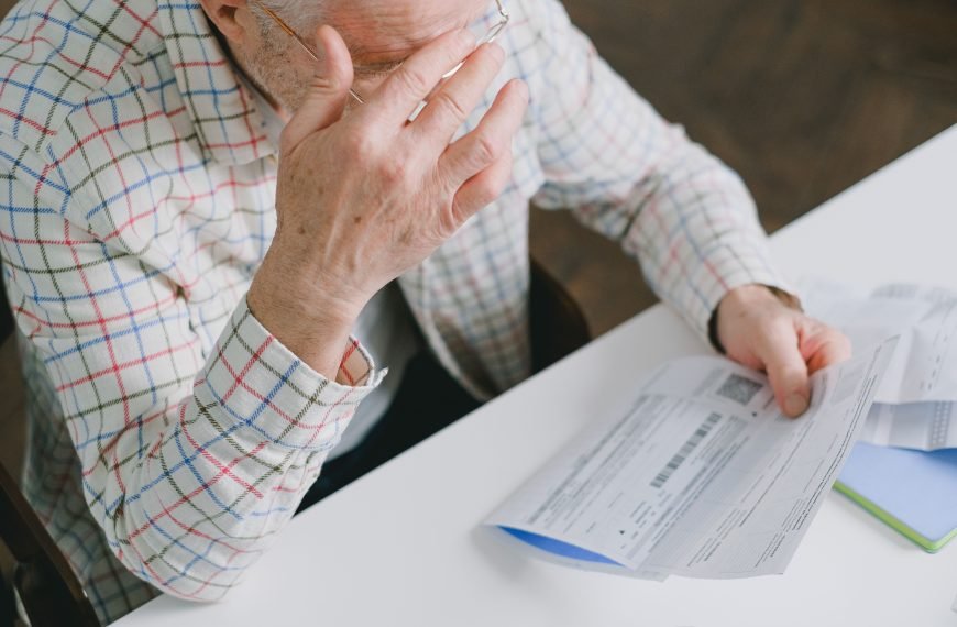 Elderly man in patterned shirt reading and holding bills at a home table, appearing focused.