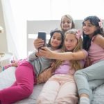 Four young girls in pajamas enjoy a joyful sleepover, capturing moments with a smartphone selfie.